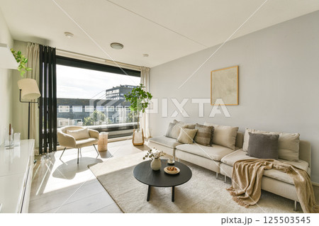 A stylish modern living room featuring a comfortable sofa, decorative elements, and large windows allowing natural light to flood the space, creating a cozy atmosphere. 125503545