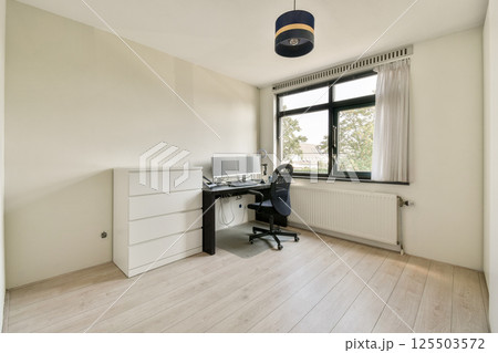 A tidy, minimalistic office featuring a desk, chair, and natural light from a large window. The space is ideal for work or study, promoting productivity and focus. 125503572