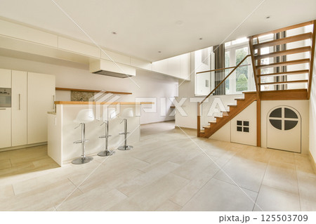A spacious and bright modern kitchen area featuring sleek design, white cabinetry, bar stools, and a wooden staircase, ensuring a stylish and functional living space. A spacious and bright modern kitchen area featuring sleek design, white cabinetry, bar stools, and a wooden staircase, ensuring a stylish and functional living space. 125503709