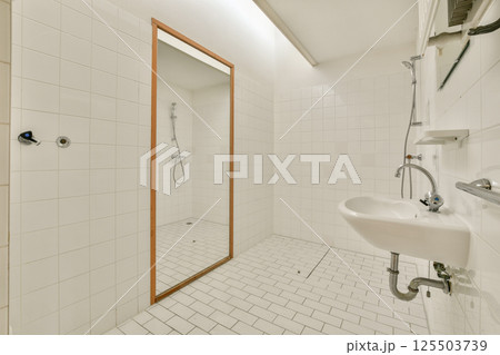 A spacious, minimalist bathroom featuring white tiled walls and floor, a large mirror, and sleek plumbing fixtures. The uncluttered design offers a clean, serene environment. 125503739