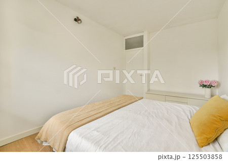 A cozy minimalist bedroom featuring a neatly made bed with soft beige linens, natural wooden floor, and simple wall decor for a calming atmosphere. 125503858