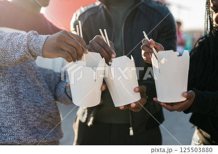 Three african american friends enjoying Asian street food outdoors on a sunny day, using chopsticks and sharing casual moments near a bright orange food truck. Warm backlight creates a cozy, friendly Three african american friends enjoying Asian street food outdoors on a sunny day, using chopsticks and sharing casual moments near a bright orange food truck. Warm backlight creates a cozy, friendly 125503880