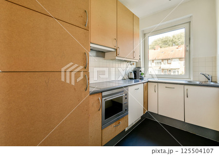 A contemporary kitchen featuring sleek wooden cabinets, a stove, and a well-lit ambiance with a window offering a glimpse of outdoor views. A contemporary kitchen featuring sleek wooden cabinets, a stove, and a well-lit ambiance with a window offering a glimpse of outdoor views. 125504107