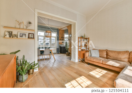A cozy modern living room featuring a brown sofa, wooden flooring, plants, and large windows that allow for plenty of natural light. 125504111