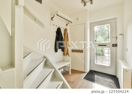 A well-lit entrance hallway with modern decor, featuring stairs, a coat rack, and a clear view of the door leading outside. 125504224