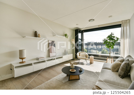 A contemporary living room featuring a sleek sofa, minimalist decor, and large windows that invite natural light, creating an inviting atmosphere. 125504265