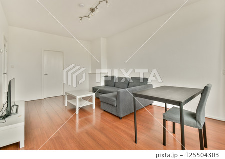 A spacious and modern living room features a grey couch, minimalist furniture, and wooden flooring, creating a stylish and inviting atmosphere for relaxation. A spacious and modern living room features a grey couch, minimalist furniture, and wooden flooring, creating a stylish and inviting atmosphere for relaxation. 125504303