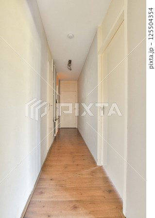 A clean and simple corridor showcasing a light-colored wall and wooden floor, leading to a closed door, creating a sense of openness and tranquility. 125504354