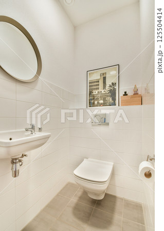 A sleek and stylish bathroom featuring a wall-mounted sink, contemporary toilet, circular mirror, and tasteful decor. The design emphasizes cleanliness and simplicity. 125504414