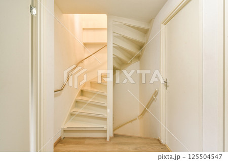 A modern interior featuring a sleek spiral staircase leading upwards. The white walls and minimalist design enhance the space's elegance. 125504547