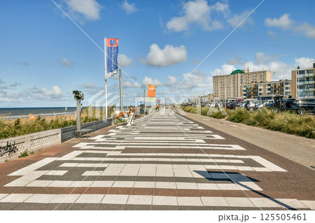 A lively coastal boardwalk with vibrant flags, walking paths, and beachside buildings under a blue sky with scattered clouds. Ideal for leisurely strolls and seaside enjoyment. A lively coastal boardwalk with vibrant flags, walking paths, and beachside buildings under a blue sky with scattered clouds. Ideal for leisurely strolls and seaside enjoyment. 125505461