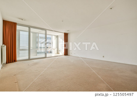 A spacious, empty living room featuring large glass windows that allow natural light. The room has light-colored walls and carpet, offering a minimalist aesthetic. A spacious, empty living room featuring large glass windows that allow natural light. The room has light-colored walls and carpet, offering a minimalist aesthetic. 125505497