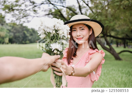 A young woman in a hat offers a bouquet of flowers, smiling warmly in a lush green park. 125508553