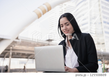 A professional woman enjoys working on her laptop outside in a modern urban setting, symbolizing business success. A professional woman enjoys working on her laptop outside in a modern urban setting, symbolizing business success. 125508609