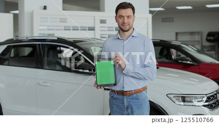 Happy man in a business suit, a manager at a car dealership holds a TabletPC with green screen in his hands and poses near the cars. 125510766