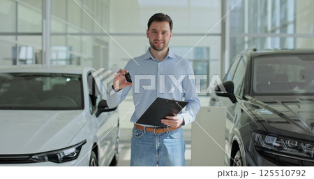 Handsome man manager at a car dealership in a business suit holding clipboard documents and car keys in his hands stands near the cars. Concept of car shopping, buying, and dealership. 125510792
