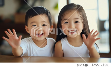 Happy siblings wave hello from a wooden table at home. Happy siblings wave hello from a wooden table at home. 125512190