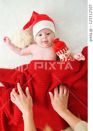 mother's hands cover a baby in a Santa's hat with a red blanket, who holds a snowman toy in his hands. mother's hands cover a baby in a Santa's hat with a red blanket, who holds a snowman toy in his hands. 125512507
