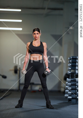 Determined woman stands dumbbells in spacious fitness studio focusing on her strength training routine enhance fitness. Modern gym equipment is visible in background. 125513681