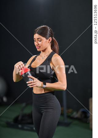 young woman in athletic wear is carefully opening shaker bottle in fitness studio. She appears focused and ready for her workout showcasing commitment health and wellness. young woman in athletic wear is carefully opening shaker bottle in fitness studio. She appears focused and ready for her workout showcasing commitment health and wellness. 125513696