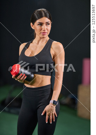 woman dressed in black athletic attire stands confidently in fitness studio while holding shaker bottle. woman dressed in black athletic attire stands confidently in fitness studio while holding shaker bottle. 125513697
