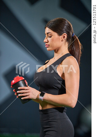 In gym environment woman dressed in sleek athletic wear stands focused while holding shaker bottle. In gym environment woman dressed in sleek athletic wear stands focused while holding shaker bottle. 125513701