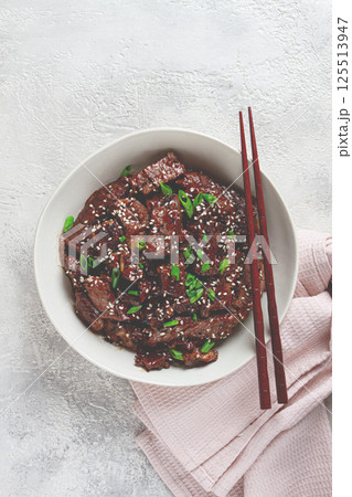 Crispy beef, with green onions and sesame seeds, in a bowl with chopsticks, on a textured surface, on a light background, no people Crispy beef, with green onions and sesame seeds, in a bowl with chopsticks, on a textured surface, on a light background, no people 125513947