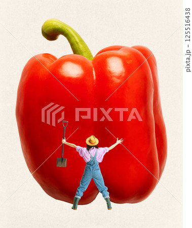 Back view of woman farmer with shovel in hand standing with wide open arms next to giant red bell pepper against light background. 125516438