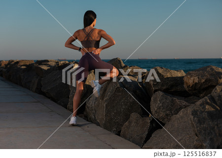 woman is stretching her back on rocky path water. She wears athletic clothing and enjoys peaceful sunset while preparing for her exercise routine. woman is stretching her back on rocky path water. She wears athletic clothing and enjoys peaceful sunset while preparing for her exercise routine. 125516837