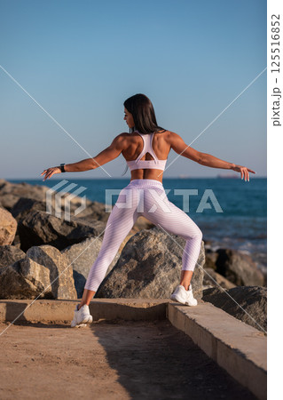 person focusing on fitness practices is seen exercising on coastal path surrounded rocks. sun sets in background creating serene atmosphere. 125516852