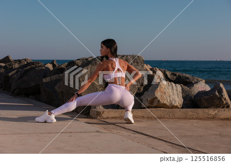 fitness enthusiast stretches on coastal pathway surrounded rocks during sunset while enjoying outdoor wellness routine. ocean is visible in background. 125516856