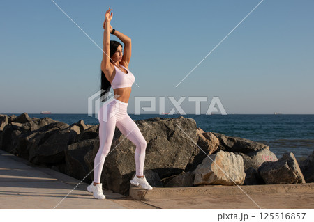 person engages in stretching exercise along rock lined beach. sun sets over water casting warm glow on fitness enthusiast dressed in light workout clothes. 125516857