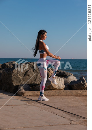 woman performs stretching exercise on coastal walkway rocks nearby. sun shines brightly as she enjoys refreshing workout ocean in background. 125516881