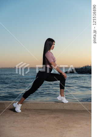 fitness enthusiast stretches lunge on coastal path as sun sets over water. serene atmosphere provides ideal setting for wellness activities. fitness enthusiast stretches lunge on coastal path as sun sets over water. serene atmosphere provides ideal setting for wellness activities. 125516890