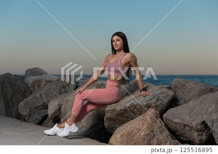 woman is confidently seated on large rocks beach during sunset wearing pink workout attire. ocean waves gently lap against shore as sun sets in background. woman is confidently seated on large rocks beach during sunset wearing pink workout attire. ocean waves gently lap against shore as sun sets in background. 125516895