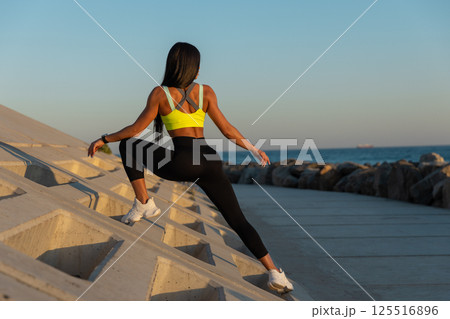 woman in bright top practices stretching exercises on rocky coastal pathway as sun sets on horizon. calming ocean creates peaceful backdrop. 125516896
