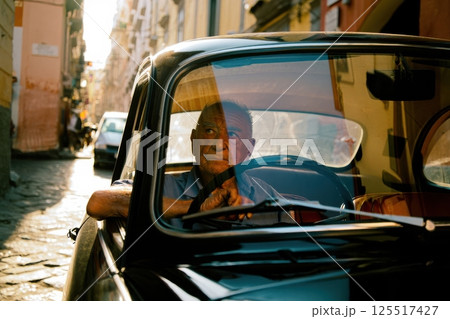 An elderly man sits in a classic car gazing thoughtfully through the windshield as warm sunlight bathes the narrow cobblestone street creating a nostalgic atmosphere. 125517427