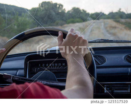 A person is gripping the steering wheel of an old car while navigating a dirt road surrounded by lush green trees and gentle hills during the late afternoon. A person is gripping the steering wheel of an old car while navigating a dirt road surrounded by lush green trees and gentle hills during the late afternoon. 125517520