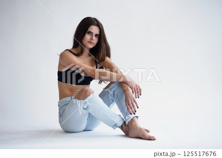 Charming skinny beautiful girl with perfect body in jeans and black top sits on white background 125517676