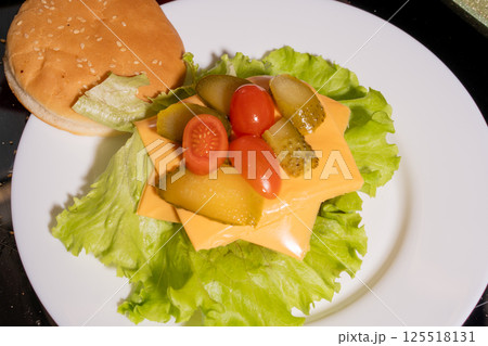 An open-faced burger with a sesame bun, lettuce, cheese, pickles, and cherry tomatoes on a white plate, highlighting vibrant burger preparation. An open-faced burger with a sesame bun, lettuce, cheese, pickles, and cherry tomatoes on a white plate, highlighting vibrant burger preparation. 125518131