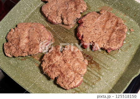 Four sizzling hamburger patties cook on a green griddle, showing texture, grease, and browning. A snapshot of burger preparation at its finest. 125518150
