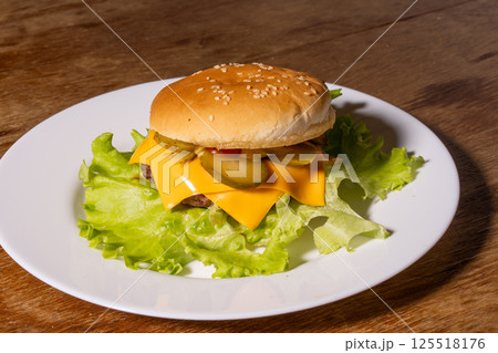 A juicy cheeseburger with a sesame seed bun, melted cheese, beef patty, and lettuce on a white plate, styled atop a rustic wooden background. A juicy cheeseburger with a sesame seed bun, melted cheese, beef patty, and lettuce on a white plate, styled atop a rustic wooden background. 125518176