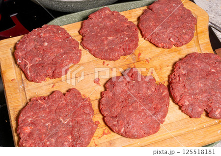 Uncooked burger patties on a wooden cutting board with hamburger buns being toasted in the background. Perfect for BBQ or homemade cooking scenes. 125518181
