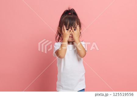 Asian little girl looks through her fingers, covers her face with both hands, scared expression, as she notices something scary or frightening, isolated on a pink background. Asian little girl looks through her fingers, covers her face with both hands, scared expression, as she notices something scary or frightening, isolated on a pink background. 125519556