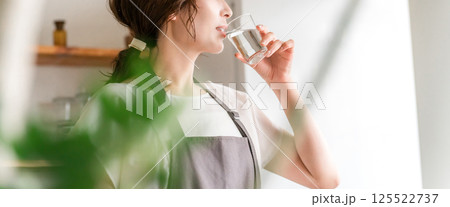 キッチンでコップで水を飲むエプロン姿の女性（水分補給,ミネラルウォーター,ウォーターサーバー） 125522737