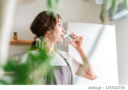 キッチンでコップで水を飲むエプロン姿の女性（水分補給,ミネラルウォーター,ウォーターサーバー） 125522738