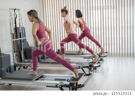 Three Asian women in pink sportswear doing pilates exercises with a reformer bed.  125523311