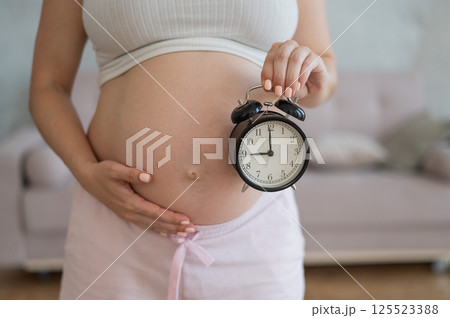 Close up of pregnant woman's belly holding alarm clock. 125523388