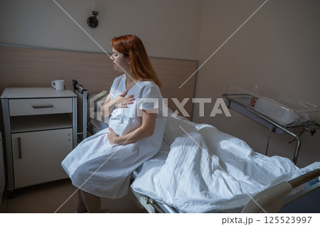 An unhappy woman sits on a couch and holds her stomach in a hospital room after giving birth. An unhappy woman sits on a couch and holds her stomach in a hospital room after giving birth. 125523997