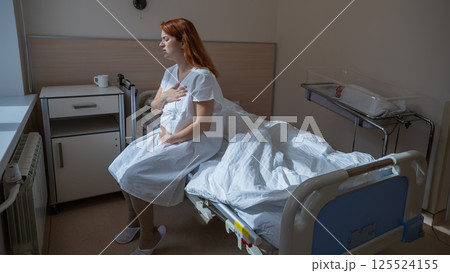 An unhappy woman sits on a couch and holds her stomach in a hospital room after giving birth. An unhappy woman sits on a couch and holds her stomach in a hospital room after giving birth. 125524155